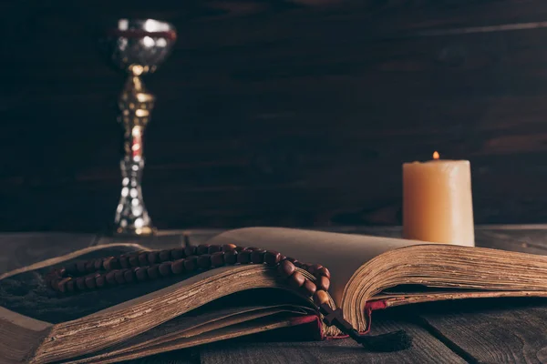 Открытая святая библия с четками, чашей и свечой на деревянном столе, Святое причастие — стоковое фото