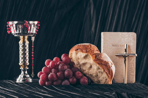 Pan, uvas, biblia, cáliz y cruces cristianas en la mesa oscura para la Santa Cena - foto de stock