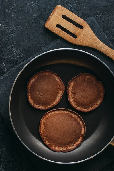 Top view of chocolate pancakes on frying pan and wooden spatula on black table — Stock Photo
