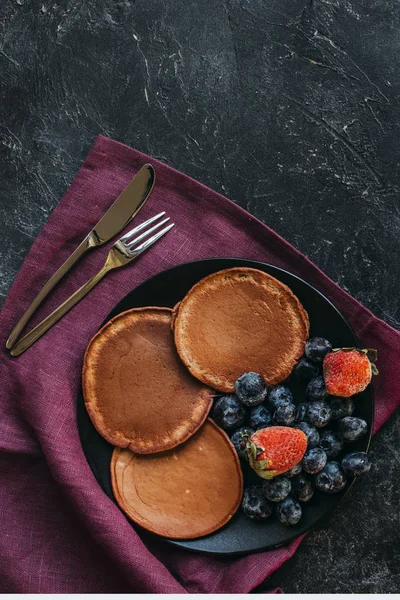 Vista superior de panquecas de chocolate com bagas e talheres na superfície de concreto preto — Fotografia de Stock