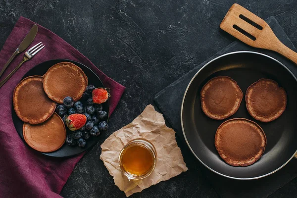 Blick von oben auf leckere Pfannkuchen auf Teller und Pfanne auf schwarzer Tischplatte — Stockfoto