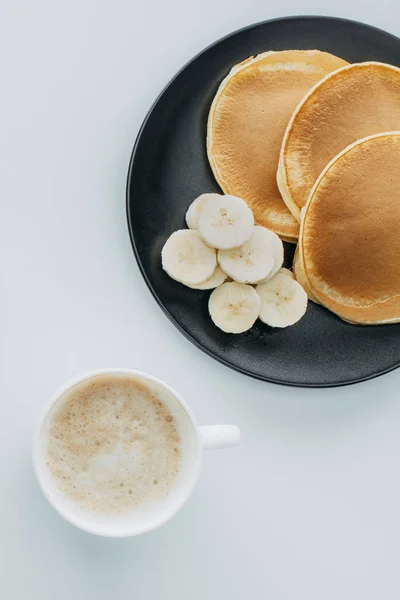 Vue de dessus des crêpes avec banane tranchée et tasse de café sur table blanche — Photo de stock