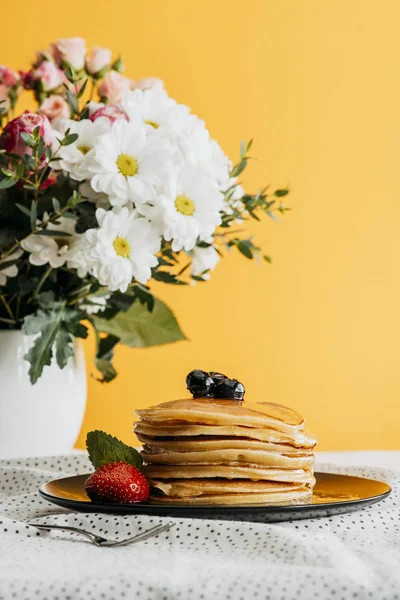 Deliziose frittelle impilate con bacche e sciroppo sul tavolo con fiori in vaso — Foto stock