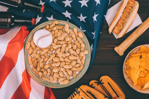 Vista superior de la bola de béisbol en el plato con cacahuetes, bate de béisbol, guante, hot dog y botellas de cerveza en la bandera americana - foto de stock