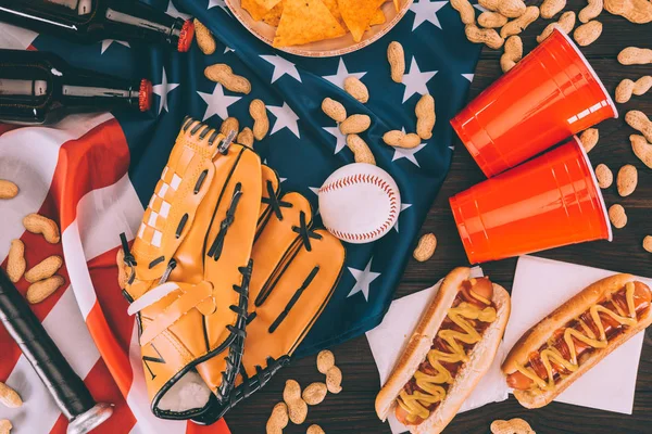 Vista superior de perros calientes, tazas de plástico, cacahuetes, botellas de cerveza, pelota de béisbol y guante con bate en la bandera americana - foto de stock