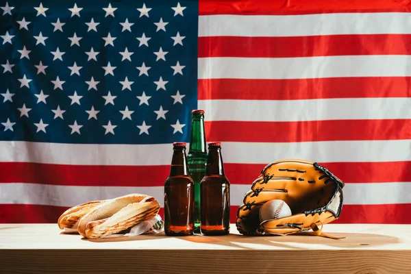 Vista ravvicinata di bottiglie di birra, hot dog, guanto in pelle e palla da baseball su tavolo in legno con bandiera dietro di noi — Foto stock