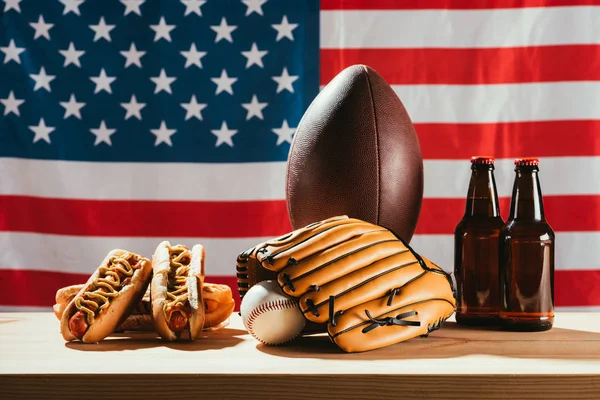 Garrafas de cerveja com cachorros-quentes e equipamentos esportivos com bandeira americana por trás — Fotografia de Stock