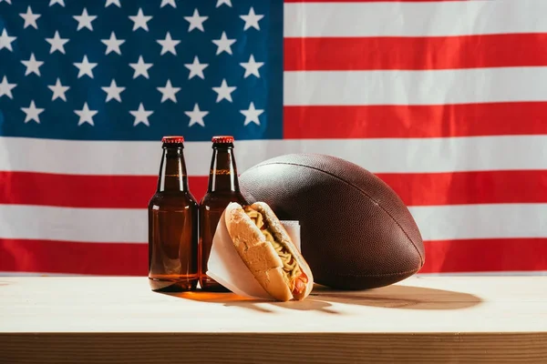 Vista ravvicinata di bottiglie di birra, hot dog e pallone da rugby sul tavolo in legno con bandiera dietro di noi — Foto stock