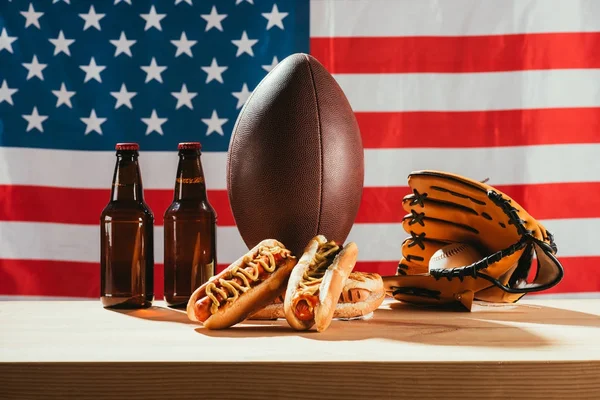 Close-up view of hot dogs, beer bottles, rugby ball and baseball glove with ball on wooden table with us flag behind — Stock Photo