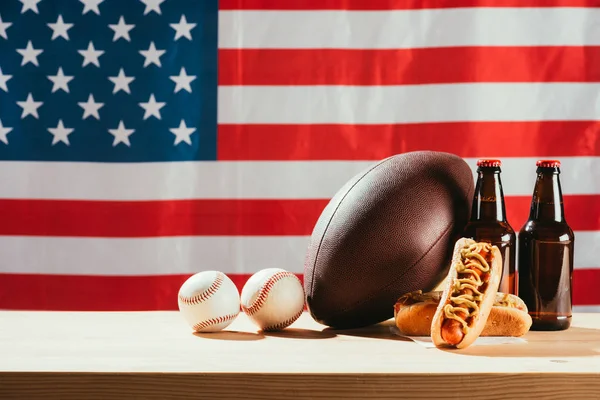 Close-up view of hot dogs, beer bottles and balls on wooden table with us flag behind — Stock Photo