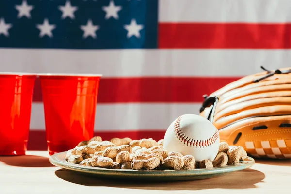 Vista close-up de bola de beisebol na placa com amendoins, copos de plástico vermelho e luva de beisebol na mesa com a bandeira da gente atrás — Fotografia de Stock