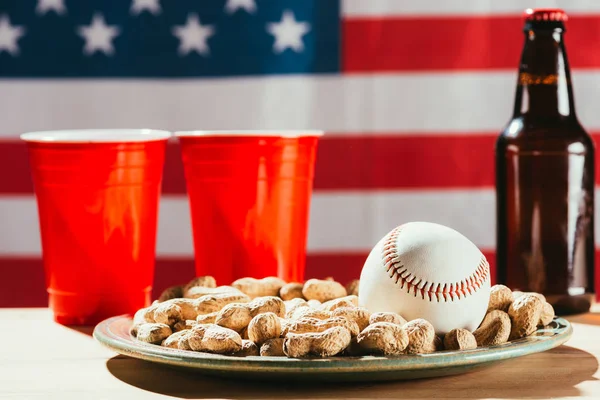 Крупным планом бейсбольный мяч на тарелке с арахисом, красными пластиковыми чашками и пивной бутылкой на столе — стоковое фото