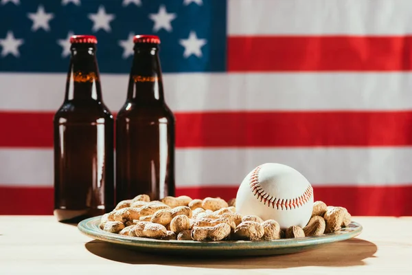 Vista ravvicinata della palla da baseball sul piatto con arachidi e bottiglie di birra con noi bandiera dietro — Foto stock