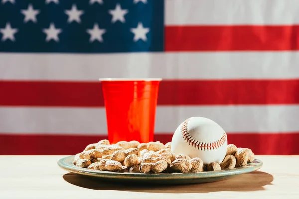 Close-up view of baseball ball on plate with peanuts, red plastic bottle and american flag behind — Stock Photo