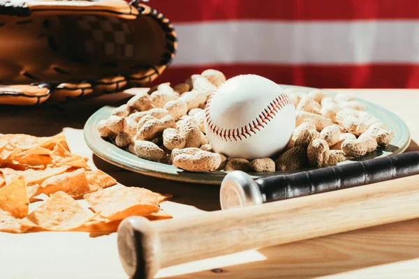 Vista de cerca de los murciélagos de béisbol, bola de béisbol en el plato con cacahuetes, bocadillos y guantes de cuero en la mesa de madera con nosotros bandera detrás - foto de stock