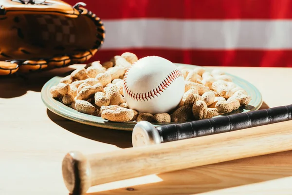 Vista close-up de bastões de beisebol, bola de beisebol na placa com amendoins e luva de couro na mesa de madeira com a bandeira atrás — Fotografia de Stock