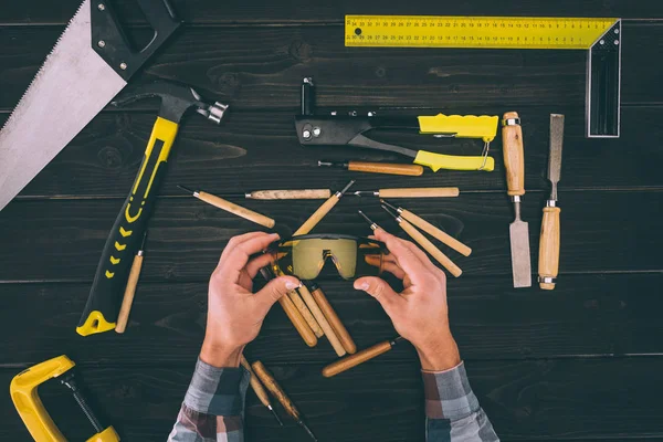 Teilansicht des Tischlers, der eine Brille mit verschiedenen Industriewerkzeugen in der Hand auf einer hölzernen Tischplatte herumhält — Stockfoto