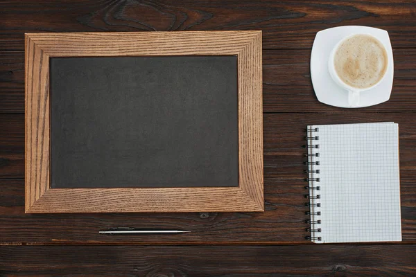 Flat lay with empty chalkboard, cup of coffee, blank notebook and pen on dark wooden tabletop — Stock Photo