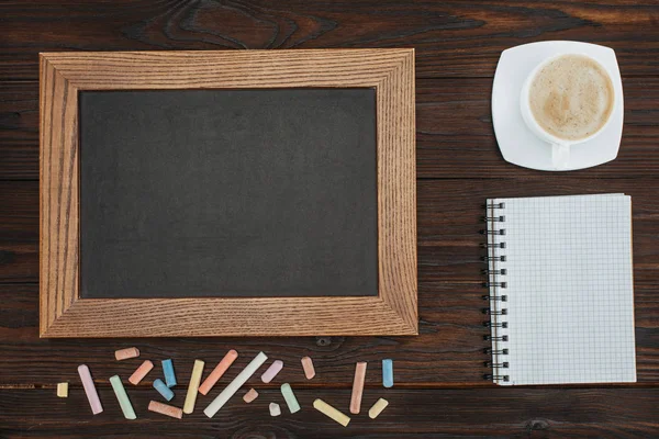 Flat lay with empty chalkboard, cup of coffee, blank notebook and chalks on dark wooden tabletop — Stock Photo