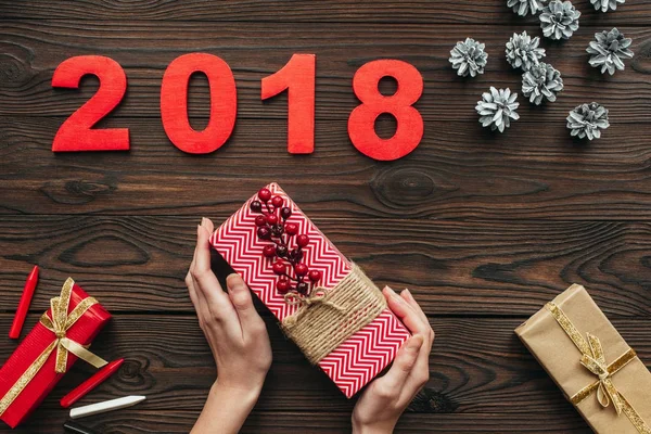 Cropped shot of woman holding wrapped gift in hands with pine cons and 2018 around on wooden tabletop — Stock Photo