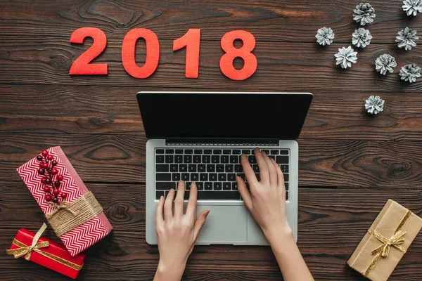 Schnappschuss von Frau beim Tippen auf Laptop mit Zahlen für 2018 und eingewickelten Geschenken auf Holztischplatte — Stockfoto