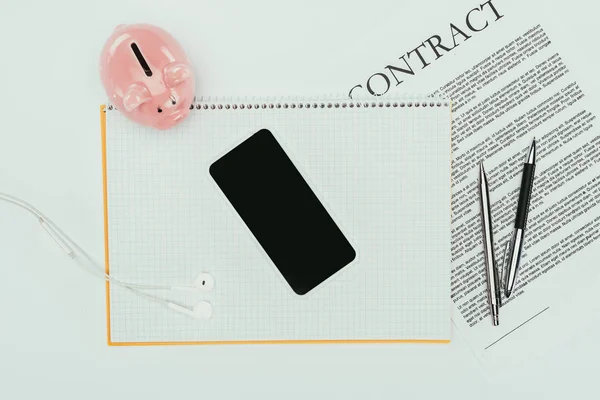 Top view of smartphone with blank screen on notebook, contract with pens and piggy bank on white — Stock Photo