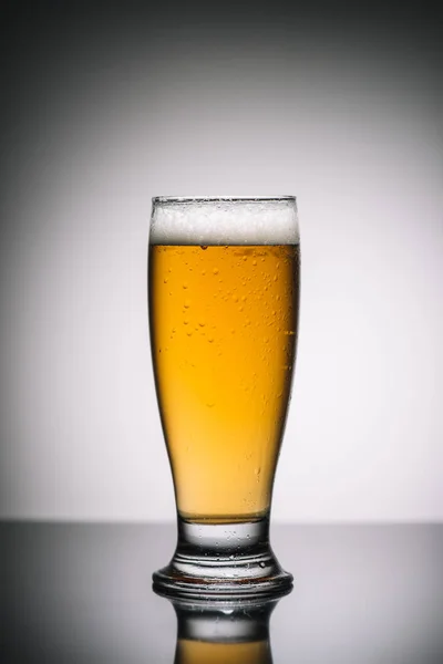 Vetro con birra leggera con schiuma sulla superficie riflettente grigia — Foto stock