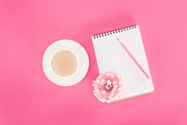 Vista superior do donut envidraçado com xícara de café e notebook em branco na superfície rosa — Fotografia de Stock