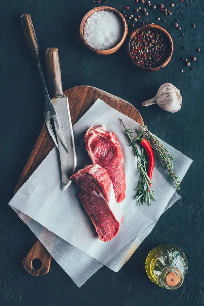 Vista superior de rebanadas de carne cruda sobre papel parchent y tabla de cortar con cubiertos - foto de stock