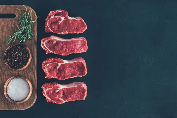 Vue du dessus des steaks crus en rangée avec des épices sur une planche en bois — Photo de stock