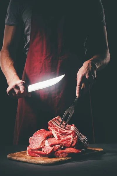 Carnicero con cuchillo y tenedor cortando carne cruda en tabla de cortar de madera - foto de stock