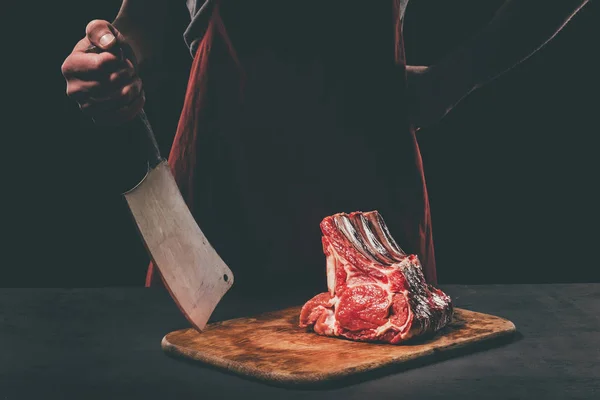 Butcher with cleaver and raw meat on wooden cutting board — Stock Photo