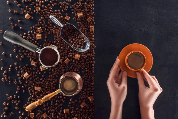 Partial top view of person holding cup of coffee and roasted coffee beans with coffee pot, scoop and sugar — Stock Photo