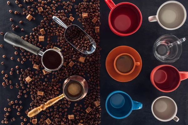 Top view of roasted coffee beans, scoop, coffee pot, coffee tamper and cup of coffee with empty cups on black — Stock Photo