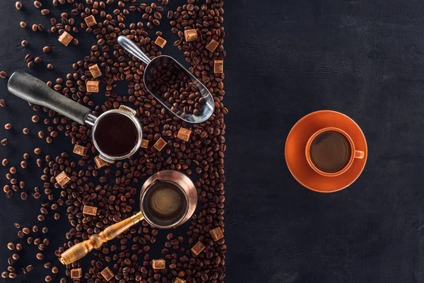 Top view of roasted coffee beans, scoop, coffee pot, coffee tamper and cup of coffee on black — Stock Photo