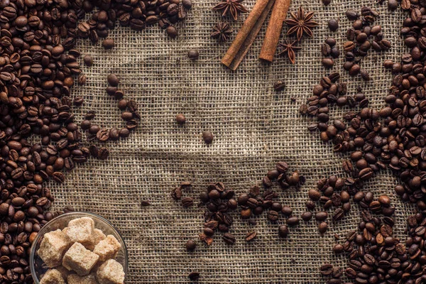 Vue du dessus des grains de café torréfiés avec bâtonnets de cannelle, anis étoilé et cassonade dans un bol en verre sur un sac — Photo de stock