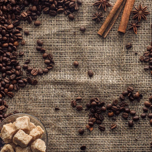 Vue du dessus des grains de café torréfiés avec bâtonnets de cannelle et sucre brun dans un bol en verre sur sac — Photo de stock