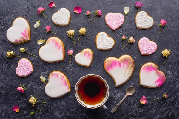 San Valentín plano con taza de té, galletas en forma de corazón acristalado y flores decorativas en la mesa oscura - foto de stock