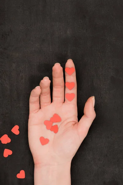 Vue partielle de la main féminine et confettis en forme de coeur rouge sur fond sombre, concept St Valentin — Photo de stock