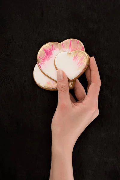 Tiro recortado de manos femeninas sosteniendo galletas en forma de corazón acristalado en el telón de fondo oscuro, San Valentín concepto de día - foto de stock