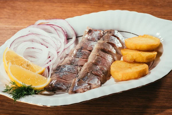 Trozos de pescado de arenque con limón, cebolla y patata en plato blanco sobre mesa de madera - foto de stock