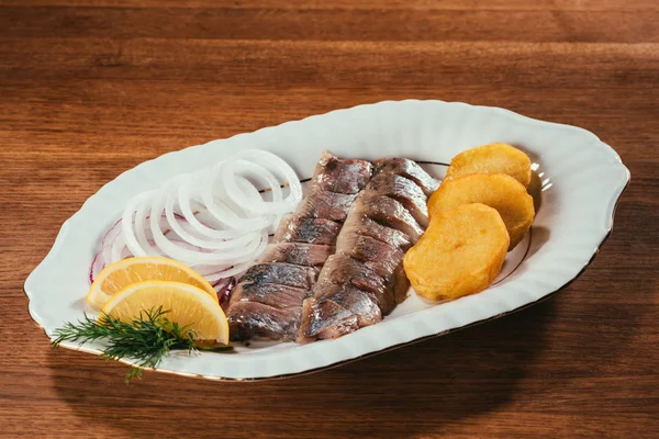 Rebanadas de pescado de arenque colocadas en el plato con cebolla y rodajas de naranja sobre la mesa de madera - foto de stock