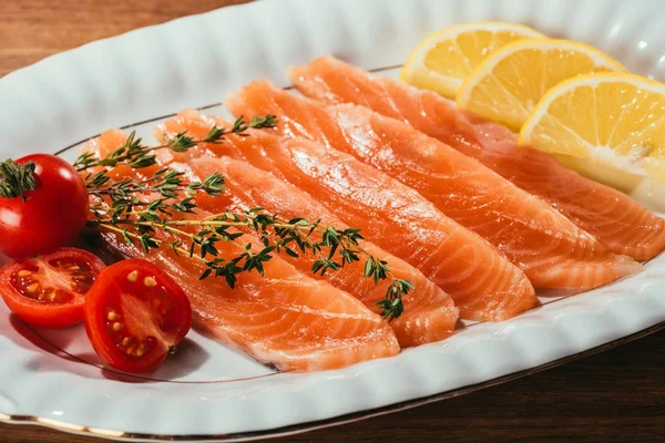 Vista de cerca de los trozos de salmón con limón, hierbas y tomates en un plato blanco - foto de stock