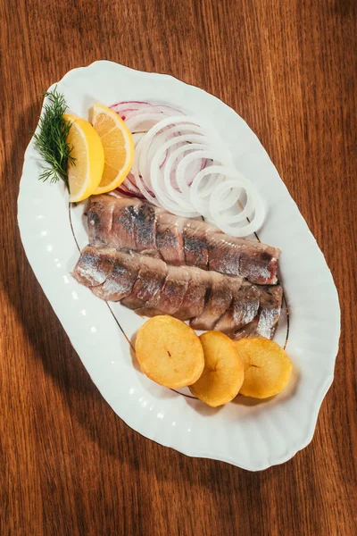 Vista superior de pedaços de peixe de arenque com limão, cebola e batata na placa branca na mesa de madeira — Fotografia de Stock