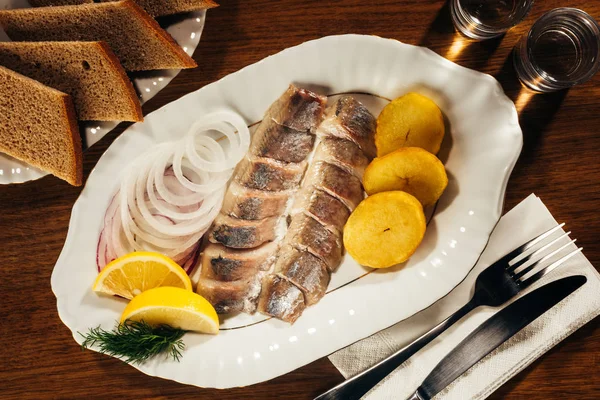 Fette di pesce aringa adagiate su piatto con fette di cipolla e arancia sopra tavolo con forchetta e coltello su tovagliolo e pane — Foto stock