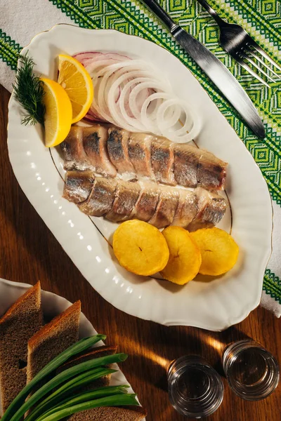 Top view of herring fish pieces with lemon and onion on white plate served with drink in glasses — Stock Photo