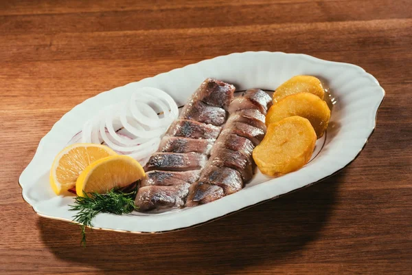 Rebanadas de pescado de arenque colocadas en el plato con cebolla y rodajas de naranja sobre la mesa de madera - foto de stock