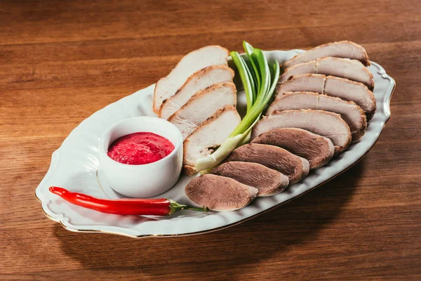 Разнообразные ломтики мяса подаются с соусом, зеленым луком и перцем на белой тарелке на деревянном столе — стоковое фото
