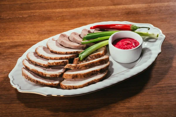 Rebanadas surtidas de carne servidas con salsa, cebolla verde y pimienta en un plato blanco sobre una mesa de madera - foto de stock