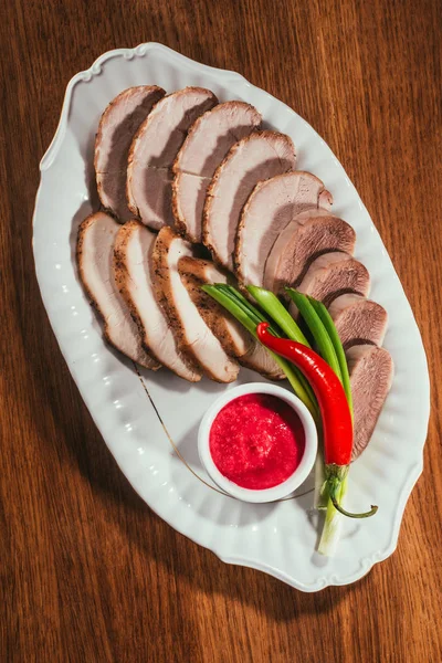 Vista superior de rebanadas surtidas de carne servidas con salsa, cebolla verde y pimienta en un plato blanco sobre una mesa de madera - foto de stock